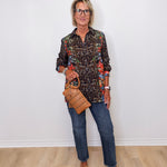 Woman wearing a floral blouse, jeans, and brown shoes, holding a woven handbag against a white wall.