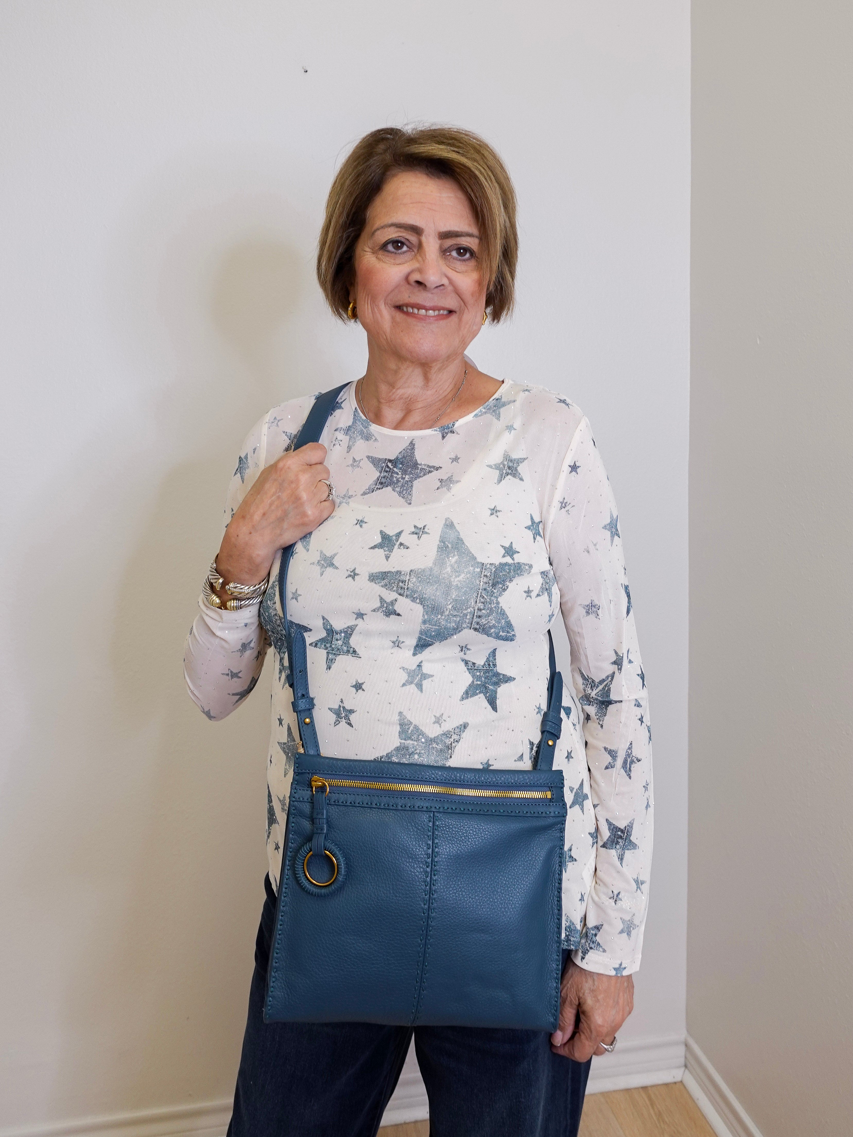 Woman wearing a star-patterned shirt and blue overalls holding a blue bag indoors.