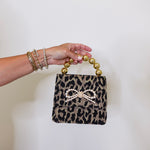 Leopard print handbag with a bow and gold handle held by a hand against a white background