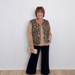 Woman wearing a leopard print vest over a beige sweater with black pants, standing against a white wall.