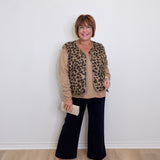 Woman wearing a leopard print vest over a beige sweater with black pants, standing against a white wall.