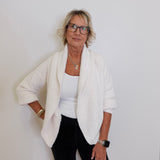 Woman wearing a white cardigan over a white top and black pants against a plain background