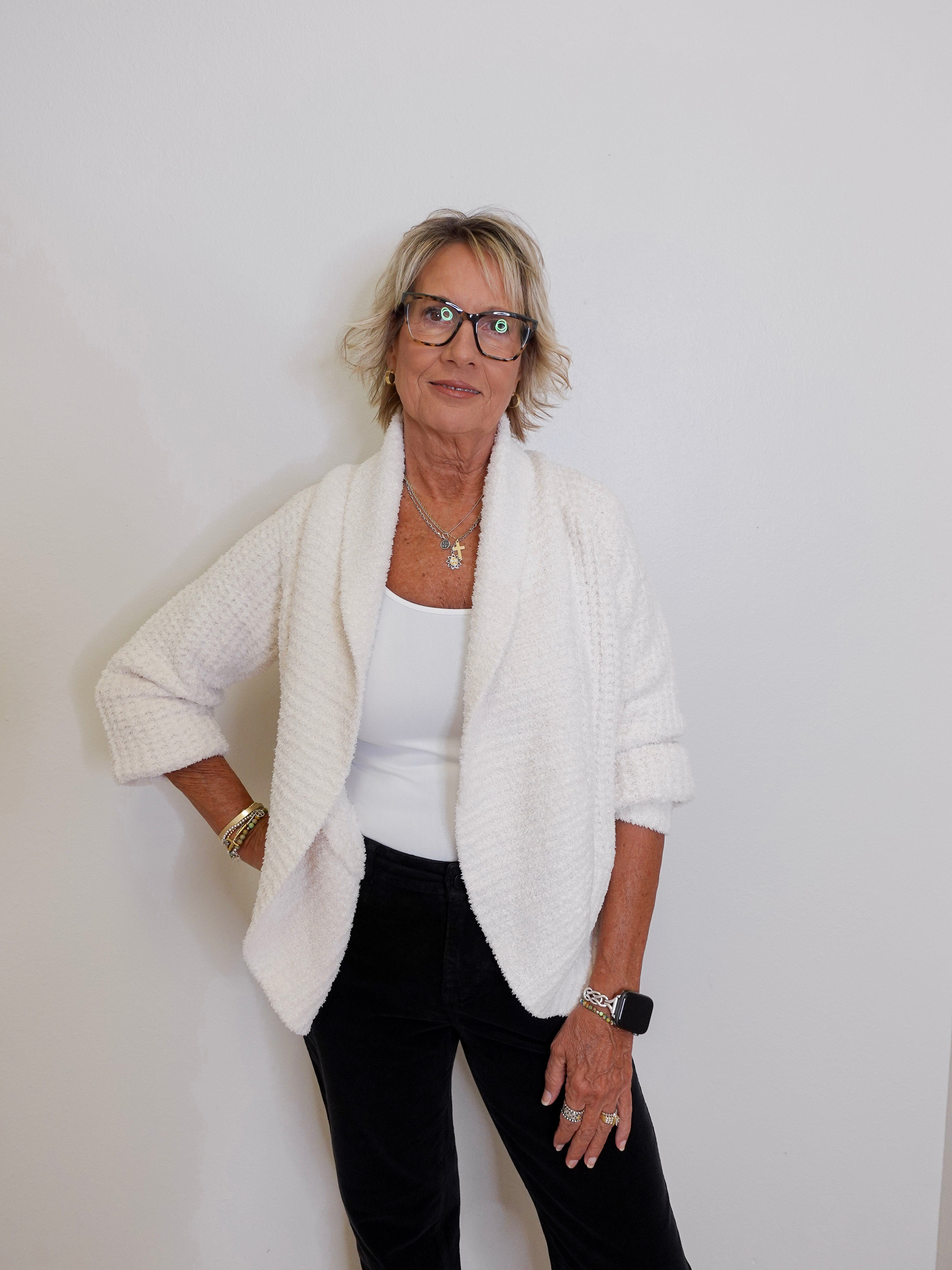 Woman wearing a white cardigan over a white top and black pants against a plain background