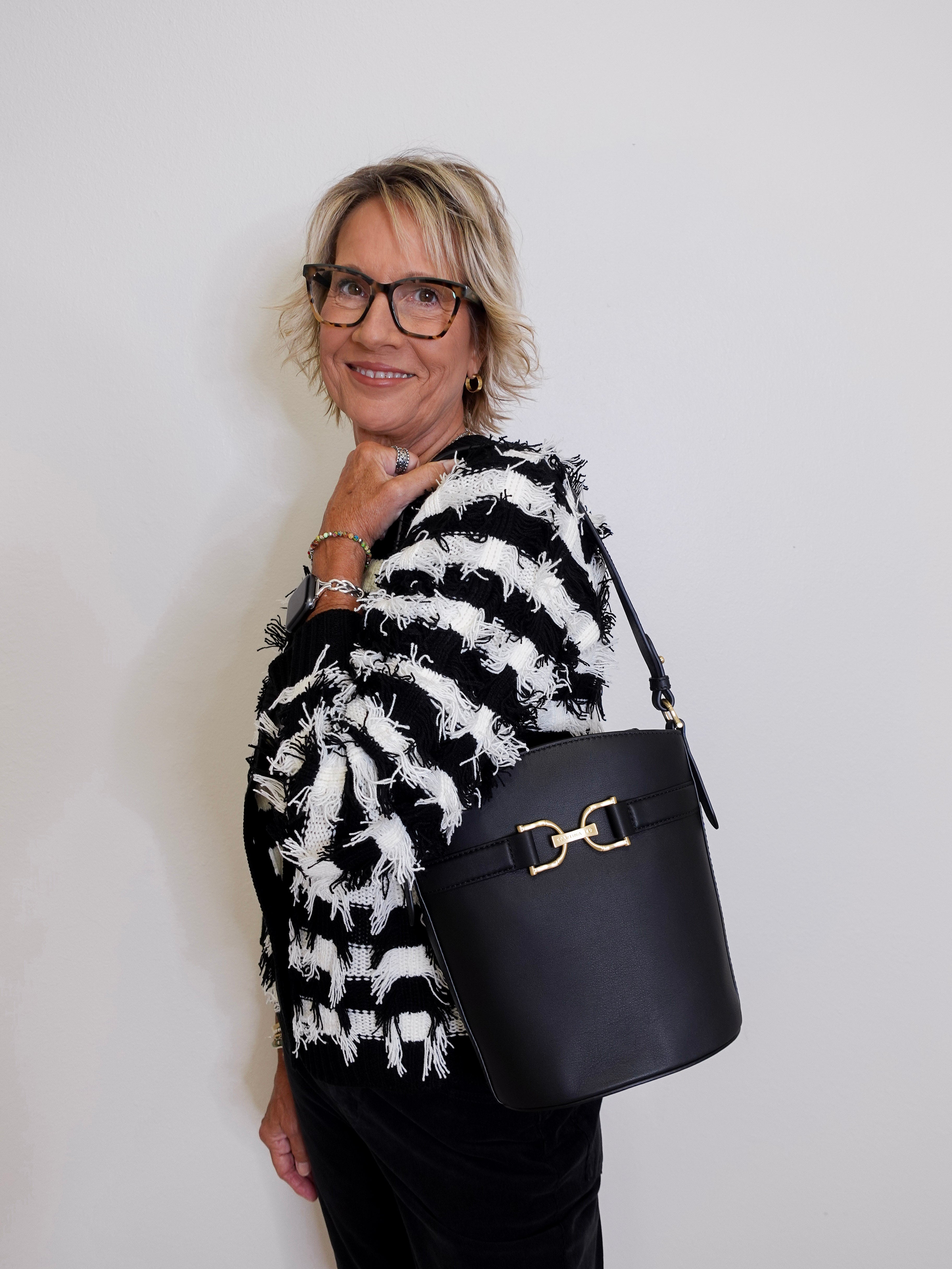 Woman wearing a black and white patterned sweater holding a black handbag against a plain background