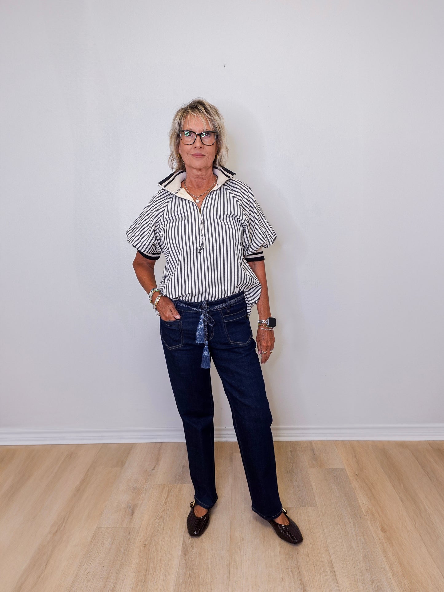 Person wearing a striped shirt and dark jeans standing against a white wall.