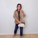 Woman wearing a beige cardigan, blue jeans, and holding a white clutch against a white wall.