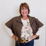 Woman wearing a brown vest over a floral shirt with a gold belt, standing against a white wall.