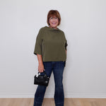 Woman wearing an olive green top and blue jeans, holding a black handbag against a white wall.