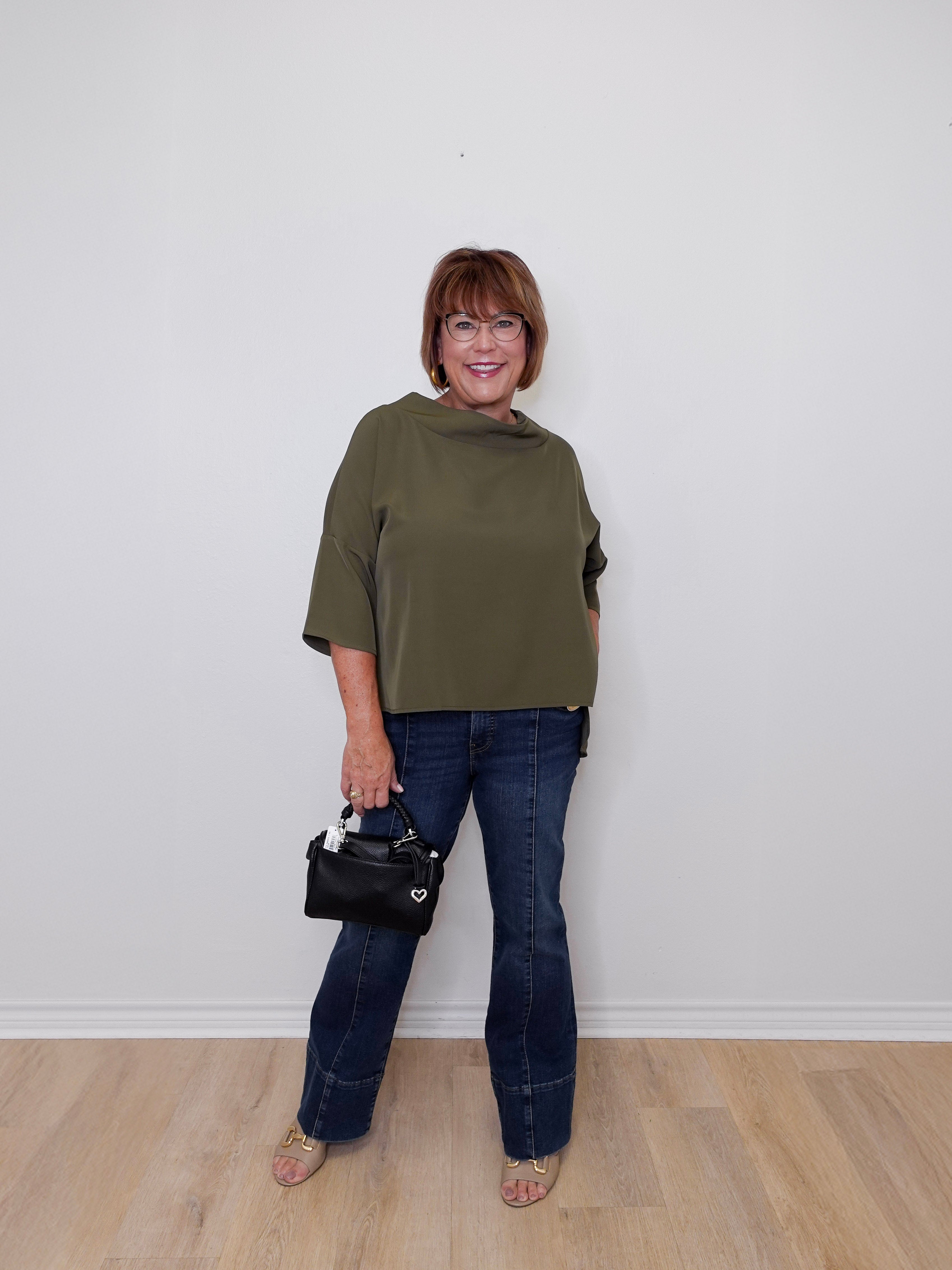 Woman wearing an olive green top and blue jeans, holding a black handbag against a white wall.