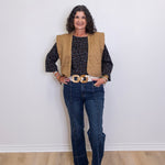 Woman wearing a beige vest, black patterned shirt, blue jeans, and brown shoes against a white wall.