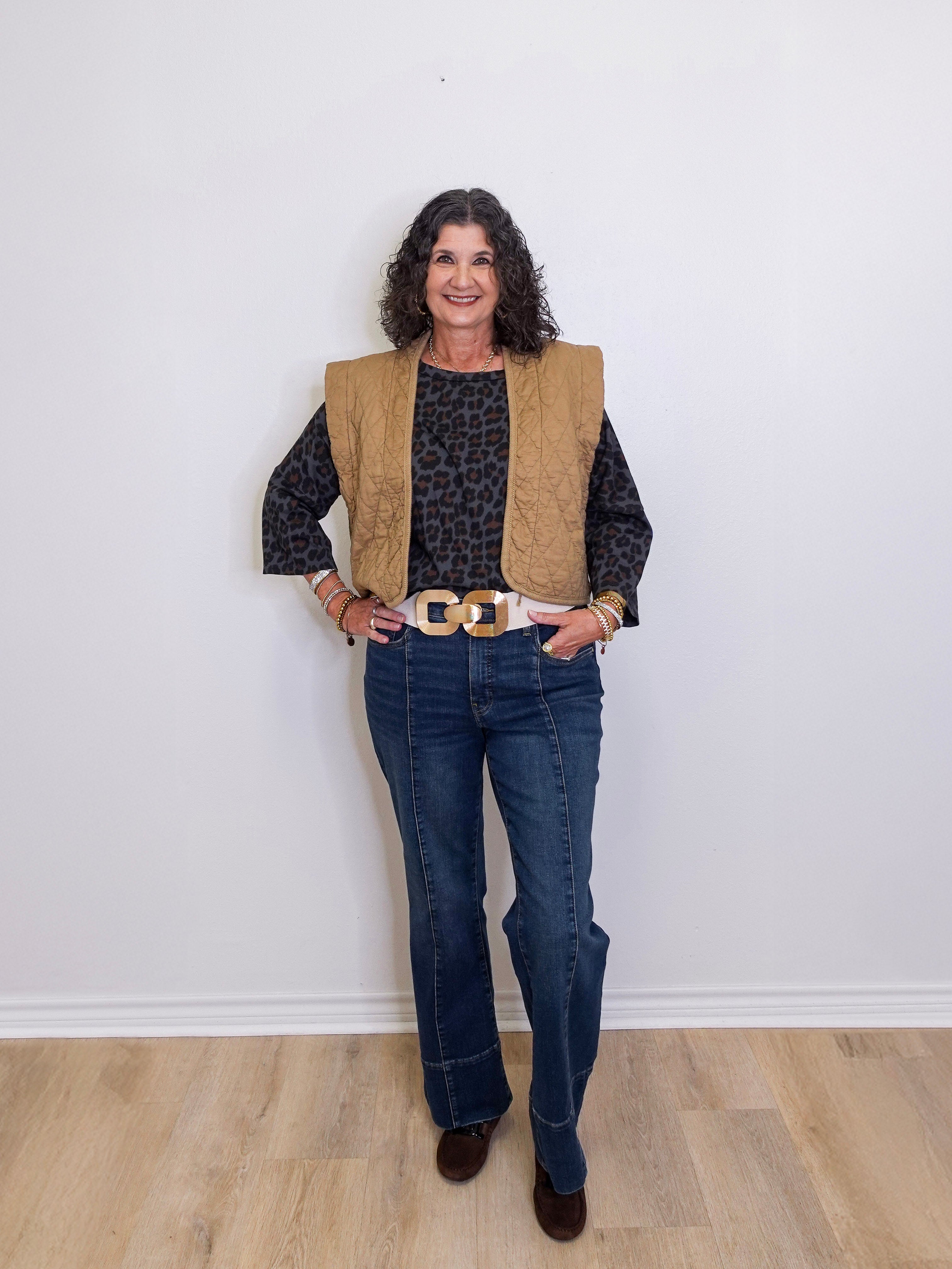 Woman wearing a beige vest, black patterned shirt, blue jeans, and brown shoes against a white wall.