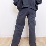 Person wearing a dark gray hoodie and pants standing against a white wall.