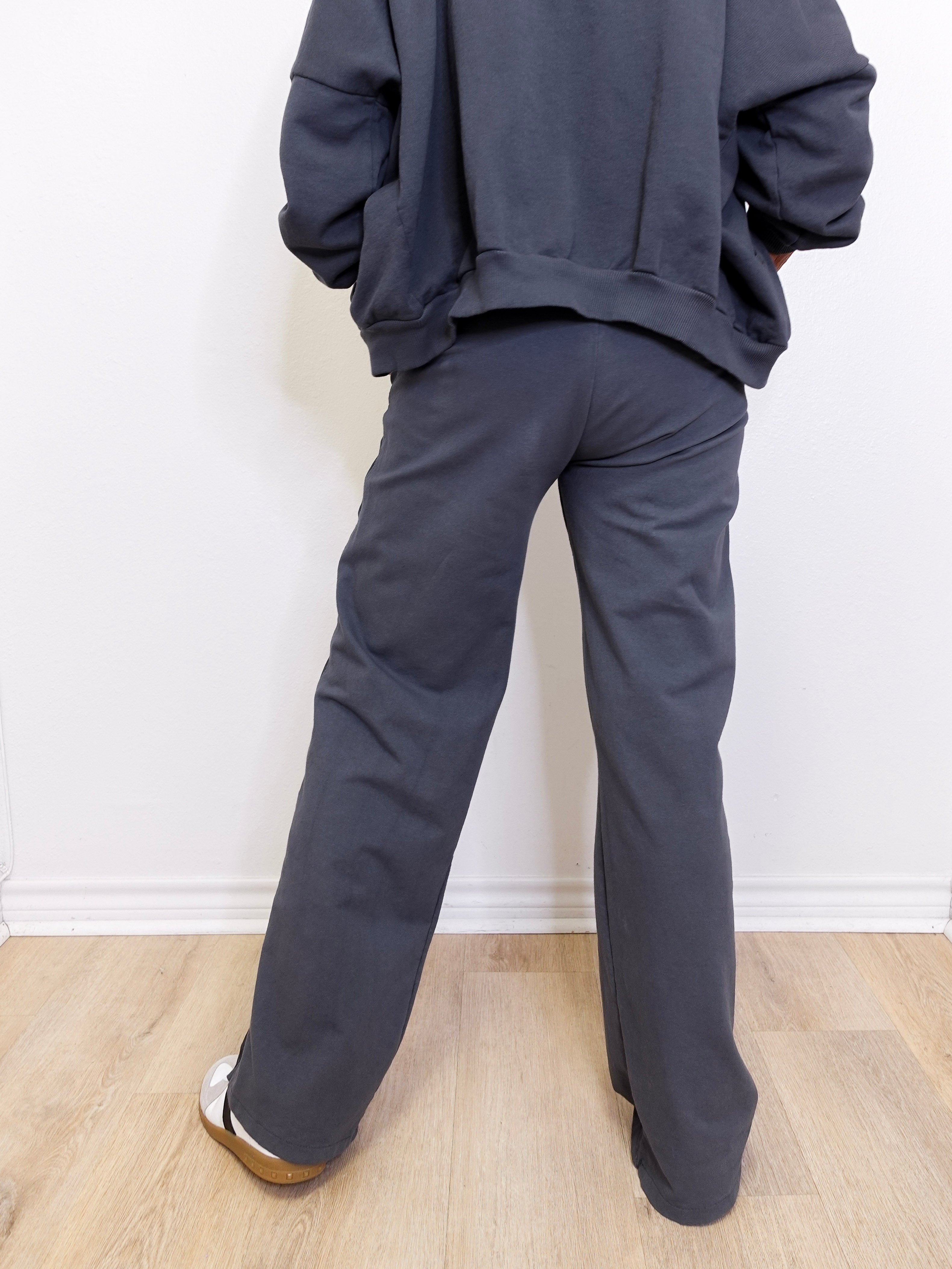 Person wearing a dark gray hoodie and pants standing against a white wall.