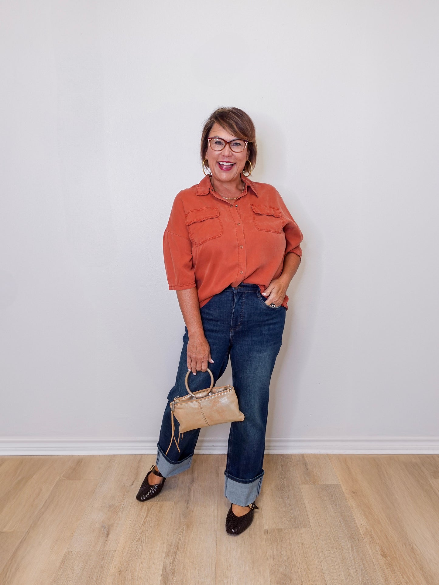 Woman wearing an orange shirt and blue jeans, holding a beige handbag against a white wall.