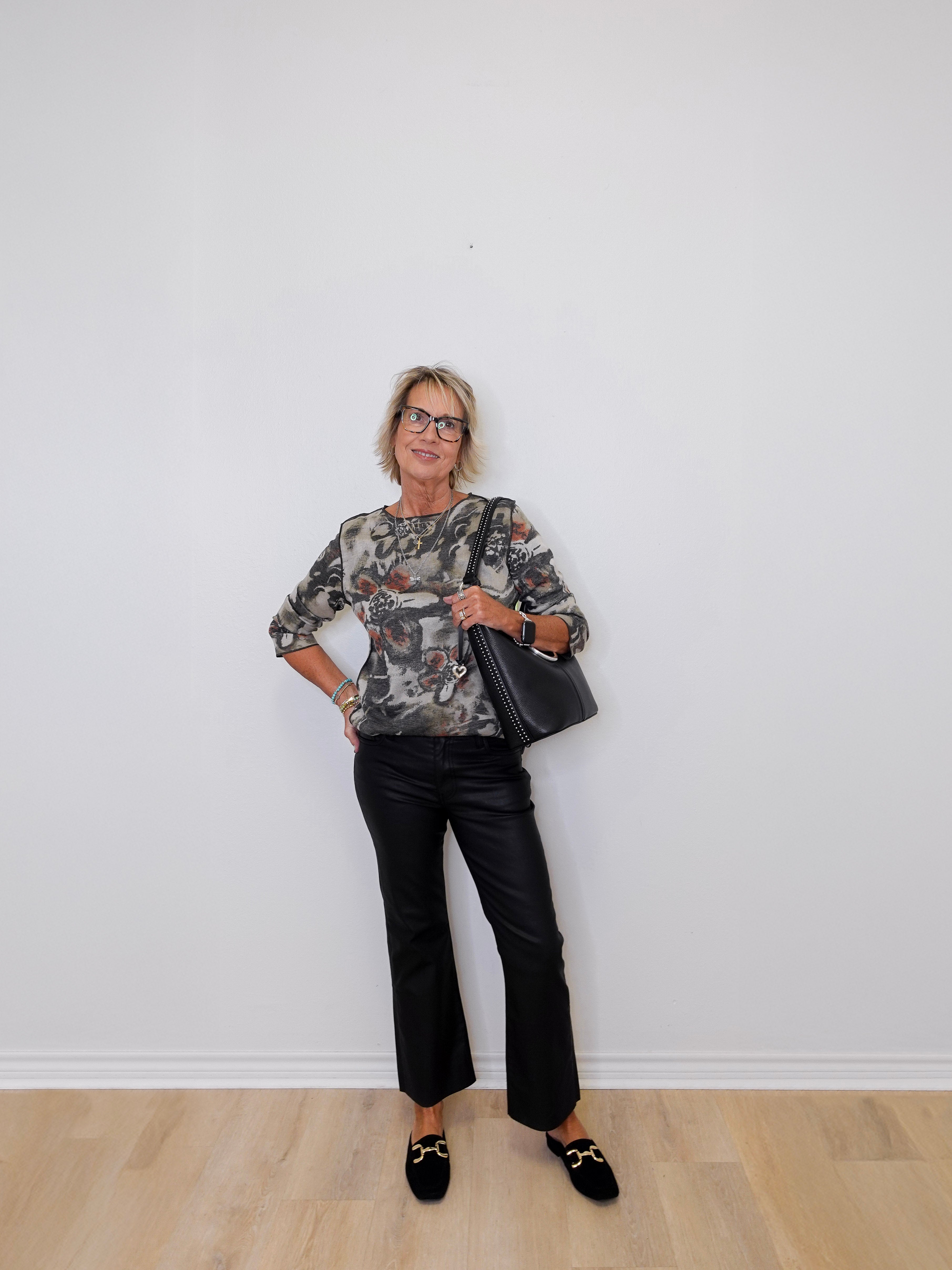 Woman in floral blouse and black pants holding a black bag against a white wall