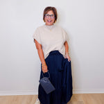 Woman wearing a beige sweater and navy skirt holding a blue handbag against a white wall.