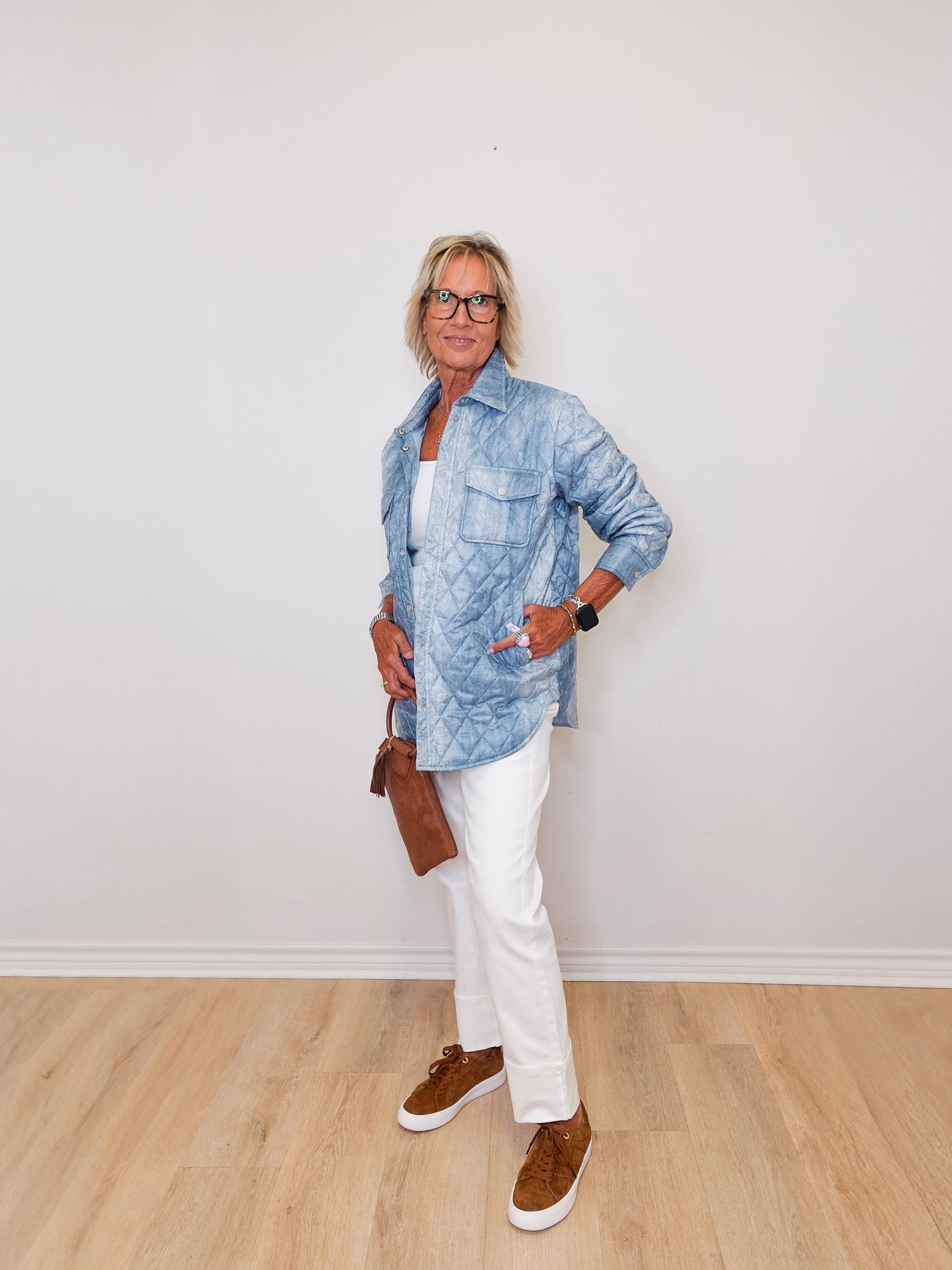 Person wearing a blue denim jacket, white pants, and brown shoes against a white wall.