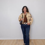 Woman wearing a floral jacket, brown top, and blue jeans against a white wall.