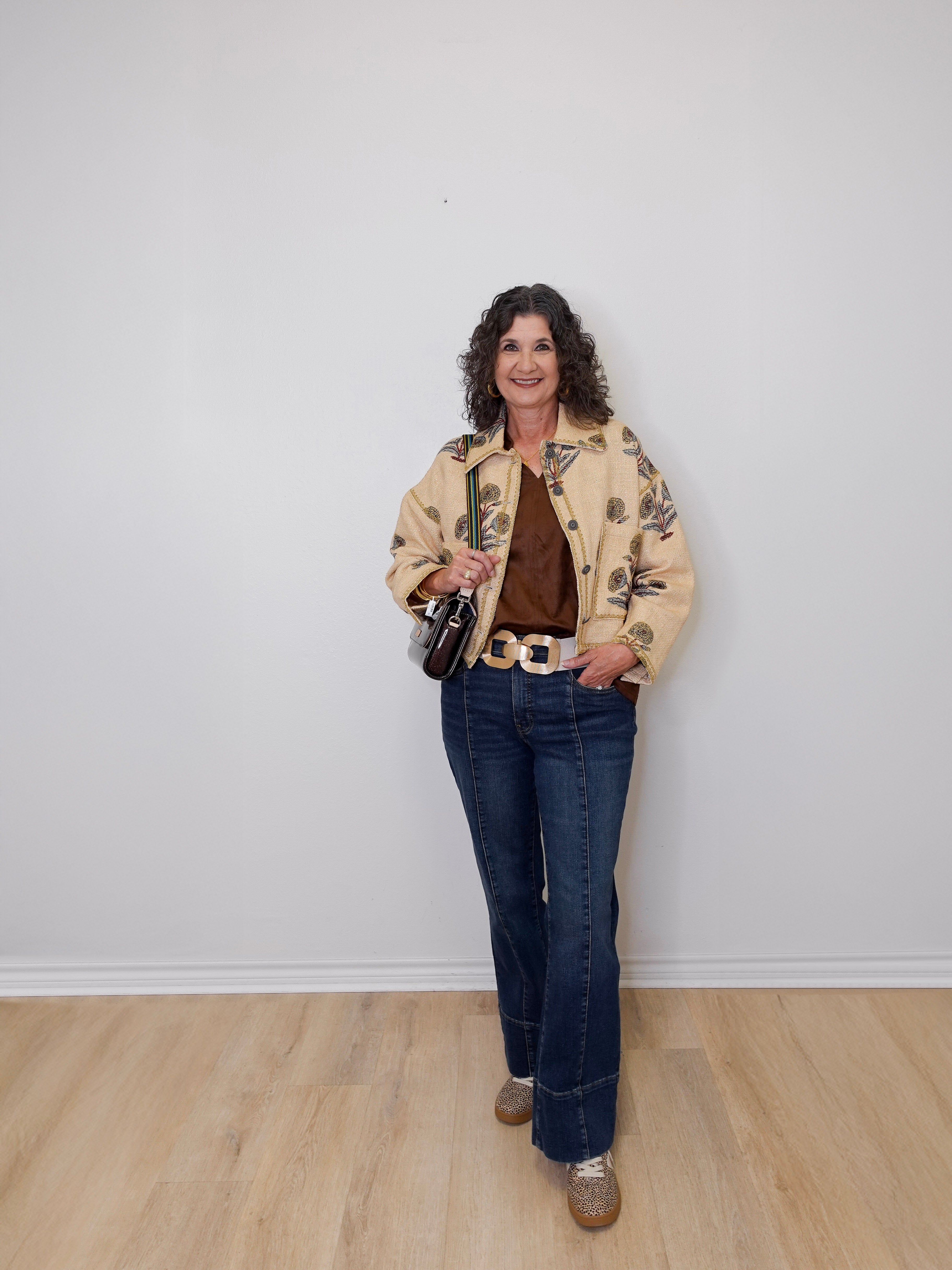 Woman wearing a floral jacket, brown top, and blue jeans against a white wall.