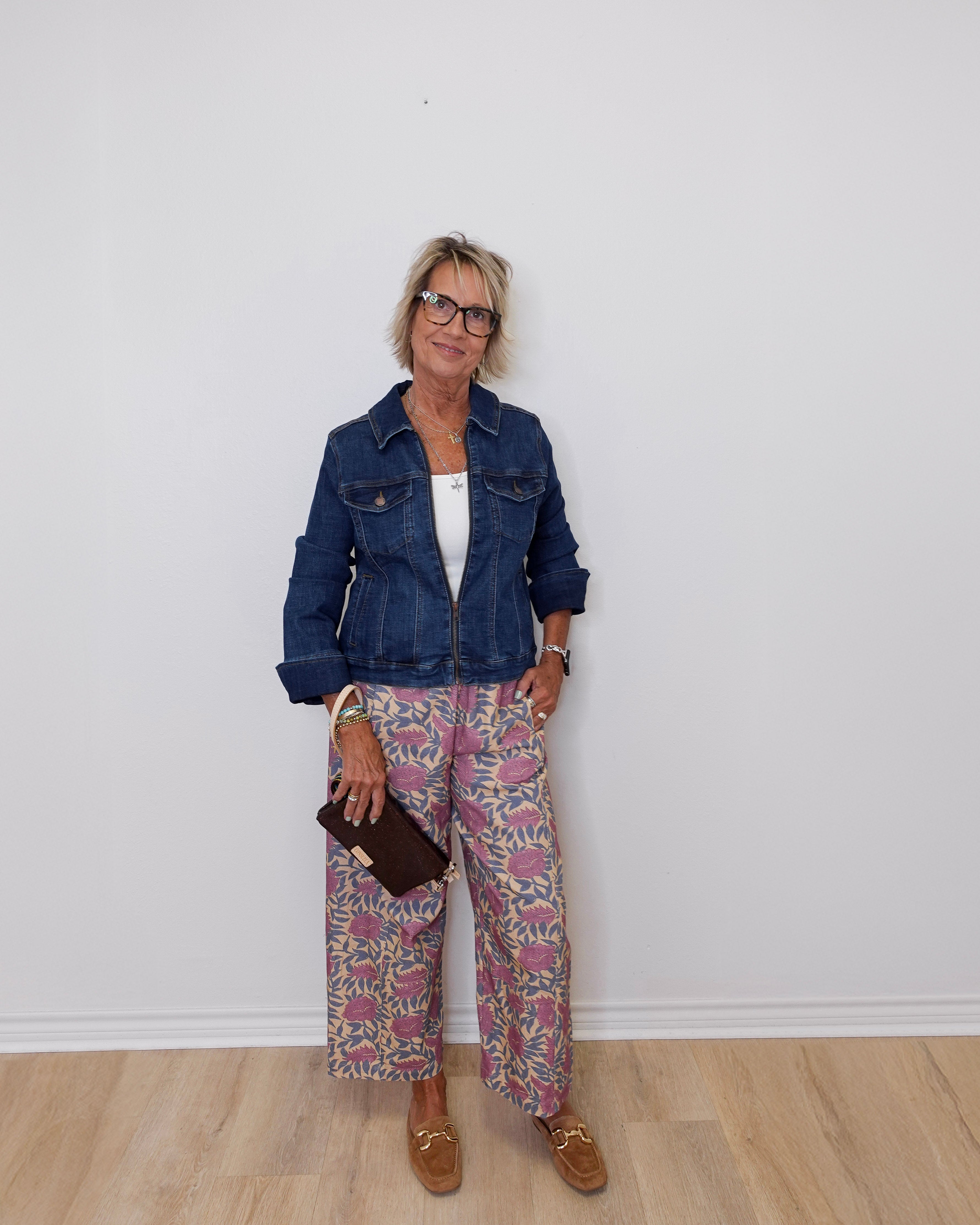 Woman wearing a denim jacket and patterned pants against a white wall.
