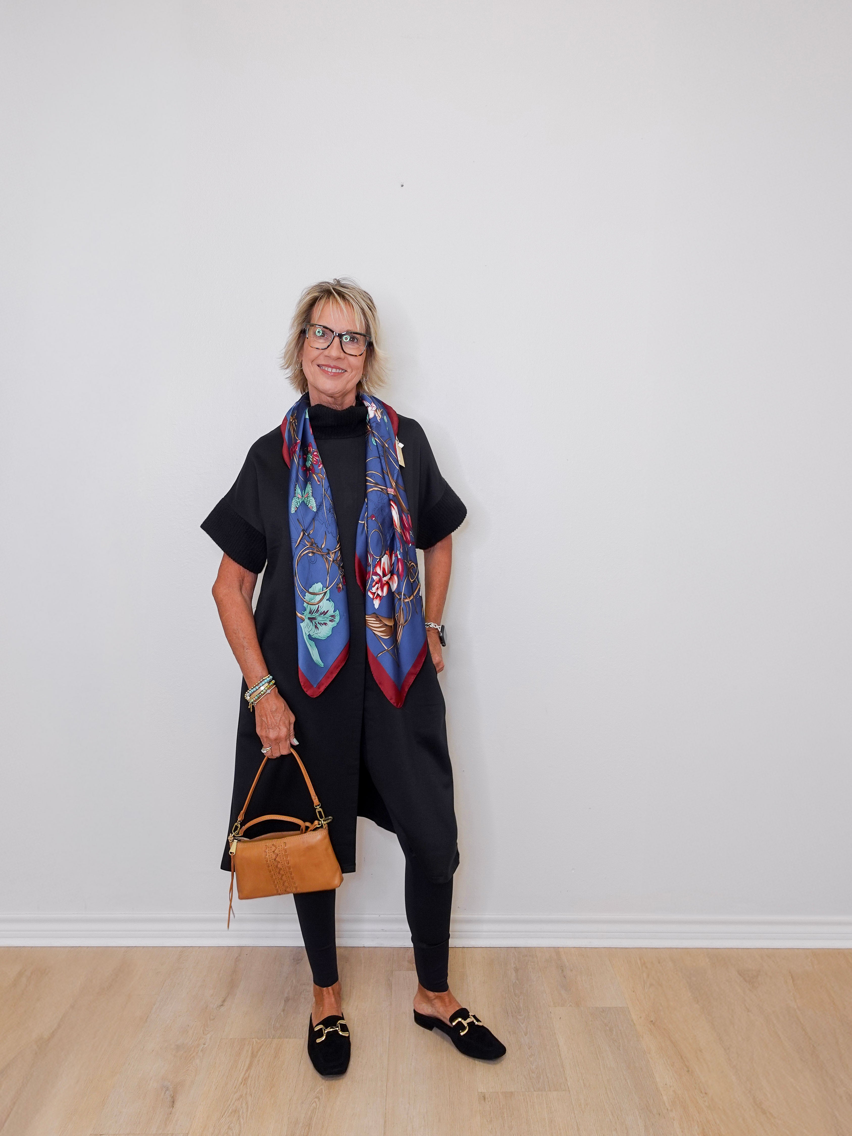 Woman wearing a colorful scarf and holding a handbag against a white wall.
