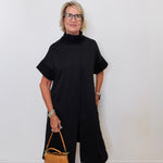 Woman wearing a black outfit holding a brown handbag against a white wall.