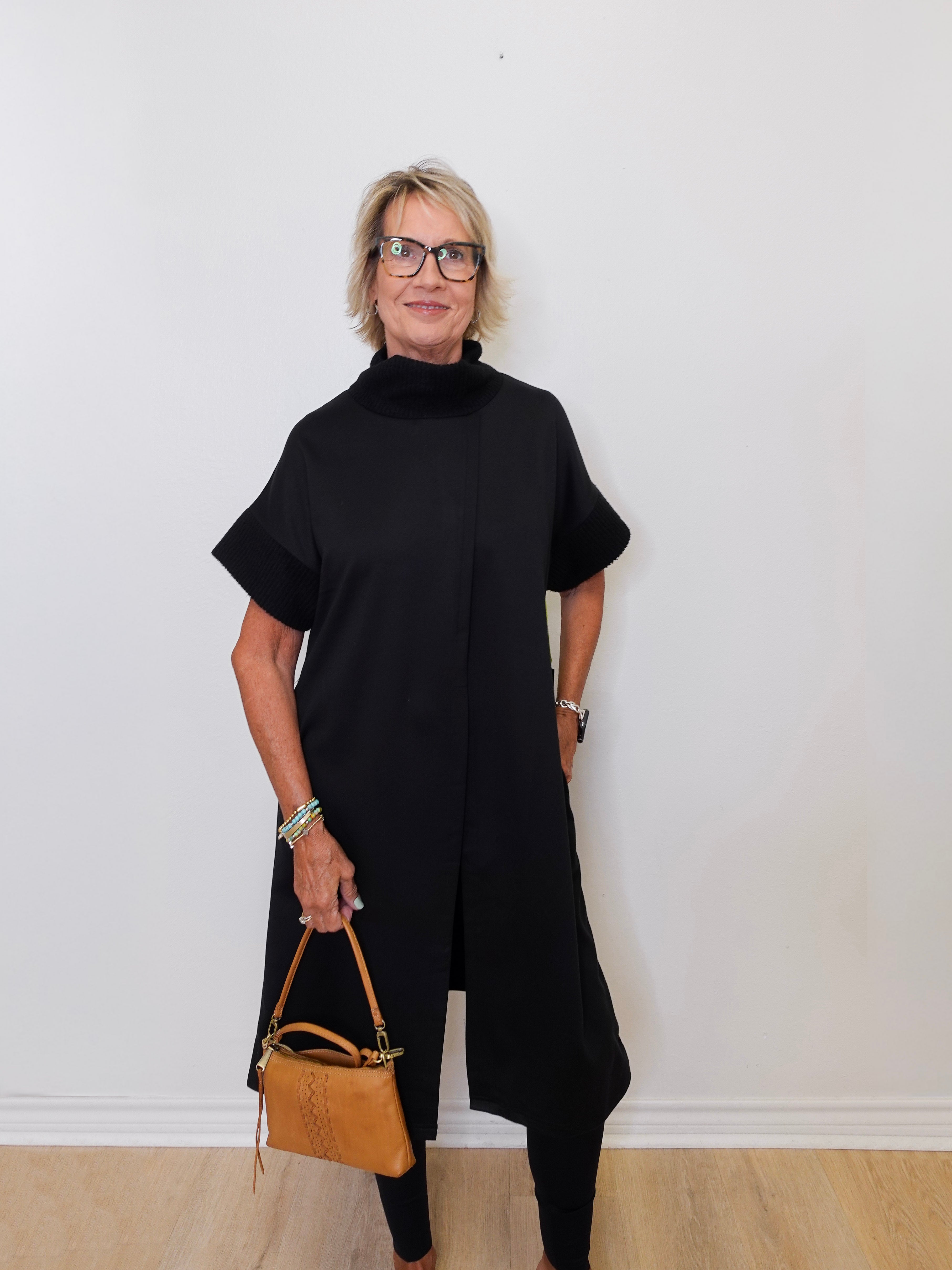 Woman wearing a black outfit holding a brown handbag against a white wall.