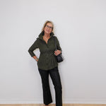 Woman wearing a green jacket and black pants against a white wall