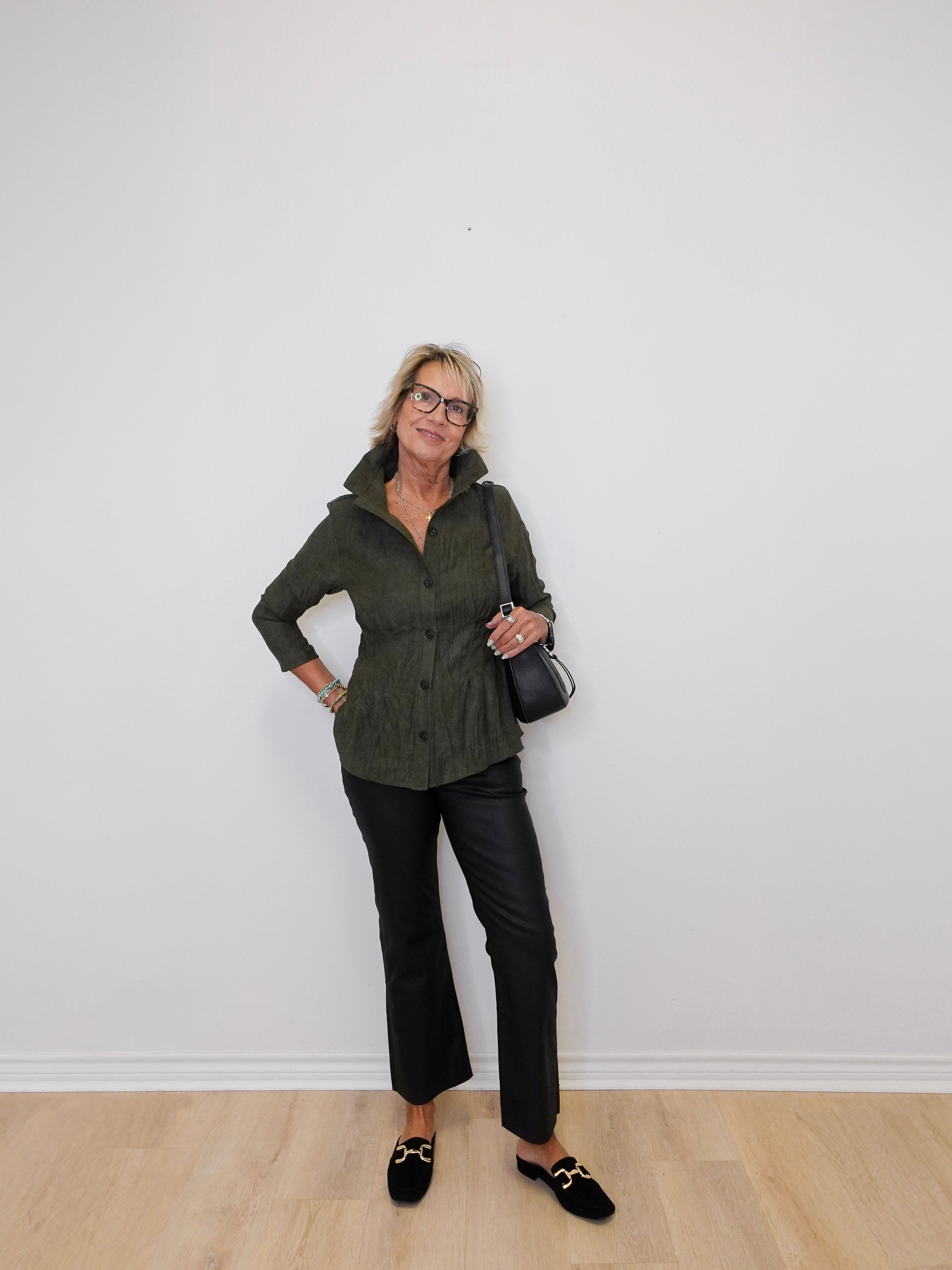 Woman wearing a green jacket and black pants against a white wall