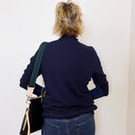 Person wearing a navy sweater and blue jeans, holding a black bag against a white wall.