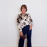 Woman wearing a patterned sweater and dark jeans against a white wall.