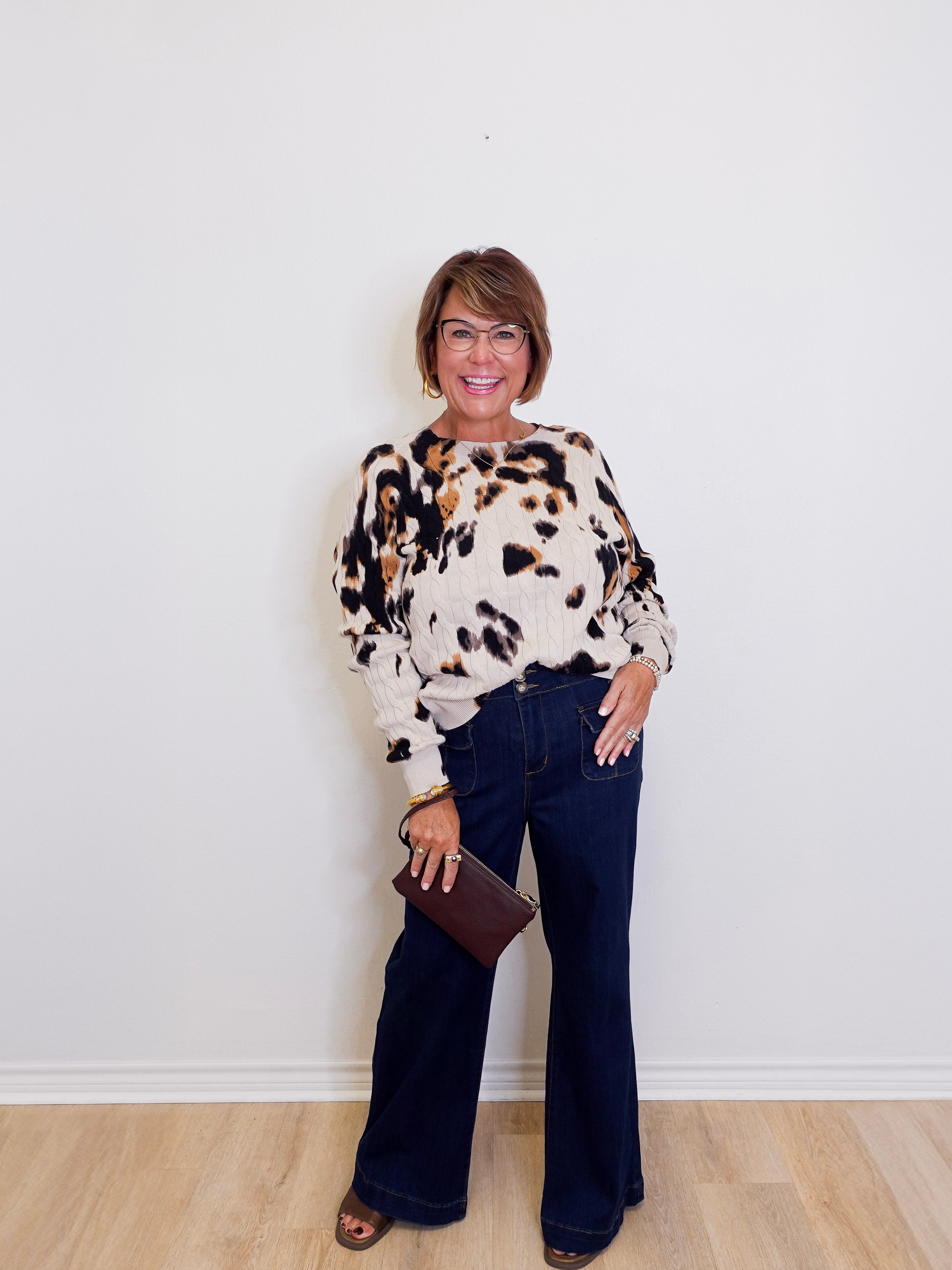 Woman wearing a patterned sweater and dark jeans against a white wall.