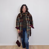Woman wearing a plaid shawl, green top, blue jeans, and white sneakers against a white wall.