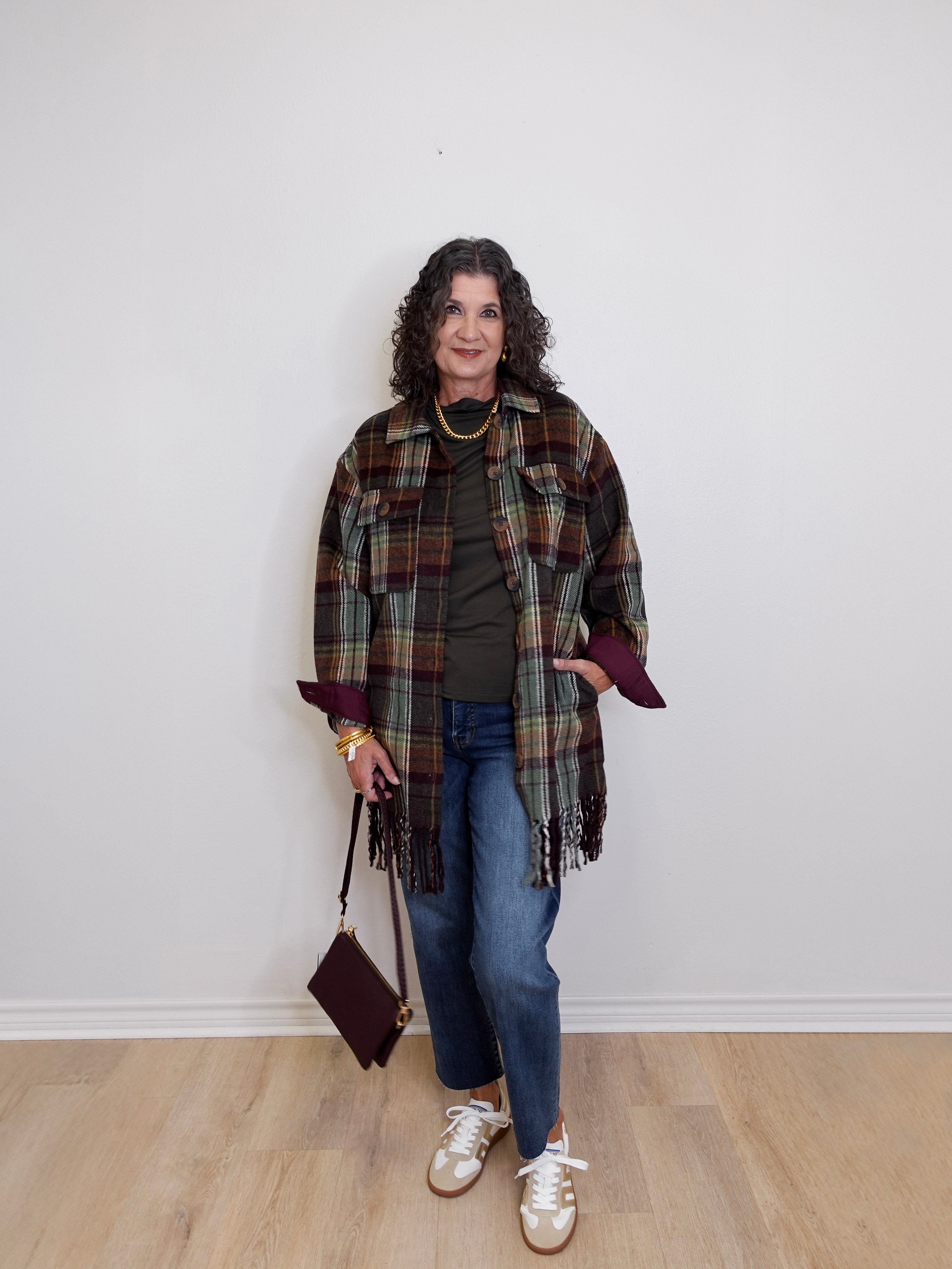 Woman wearing a plaid shawl, green top, blue jeans, and white sneakers against a white wall.