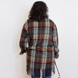Person wearing a plaid jacket with fringe, standing against a white wall.