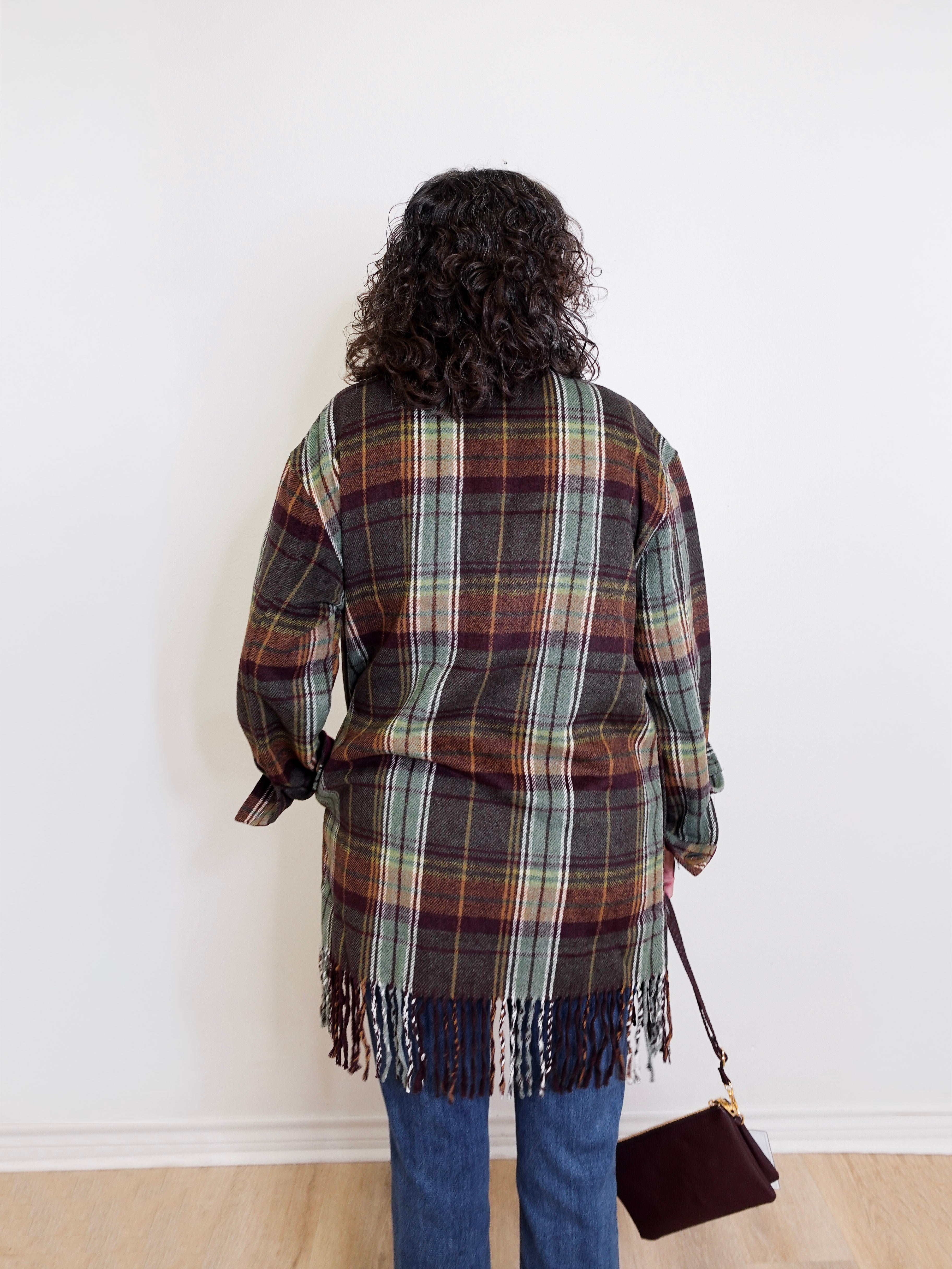 Person wearing a plaid jacket with fringe, standing against a white wall.