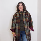 Woman wearing a plaid shawl over a green top and blue jeans, standing against a white wall.