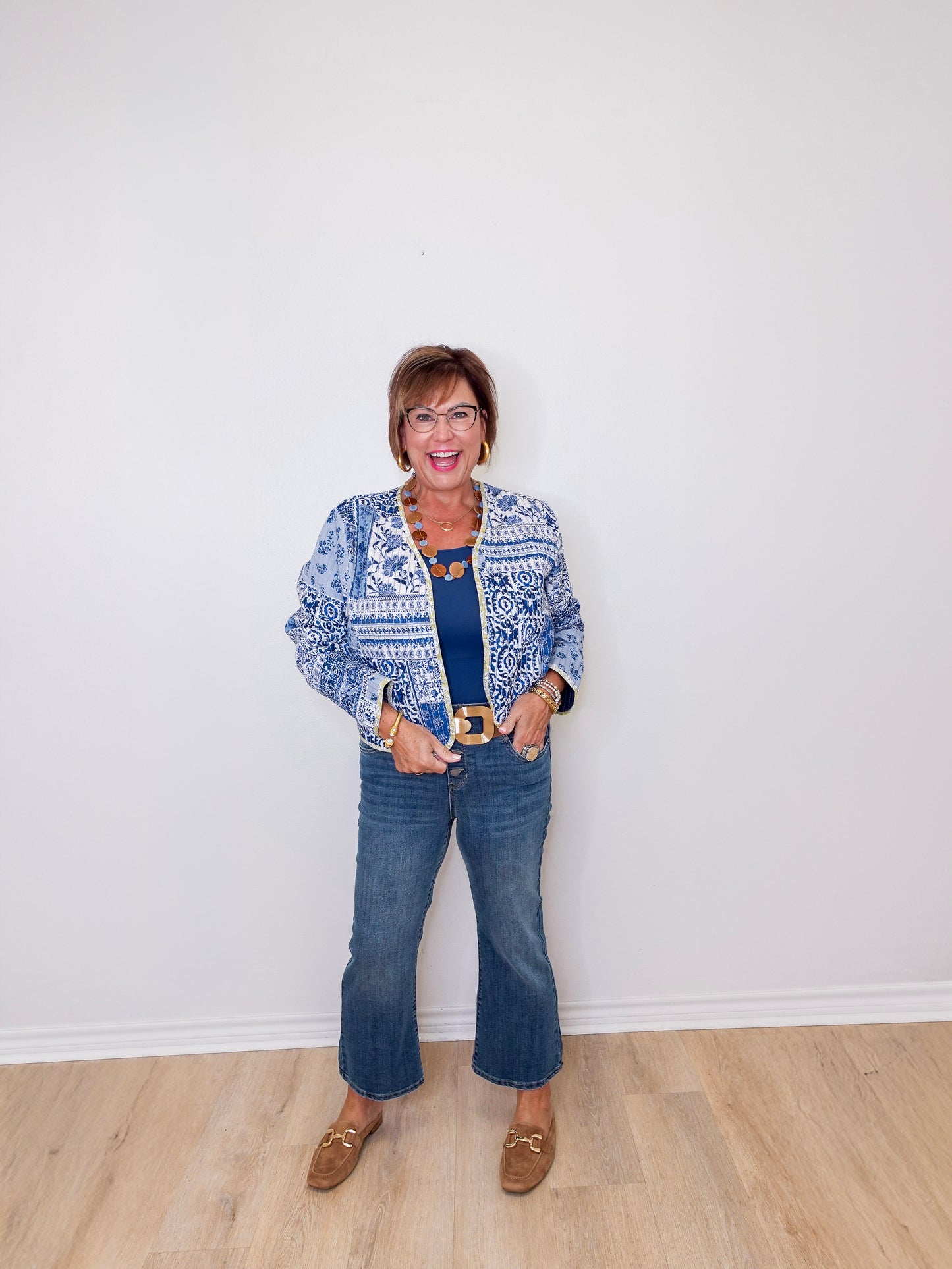 Woman wearing a blue patterned jacket, blue jeans, and brown shoes against a white wall.