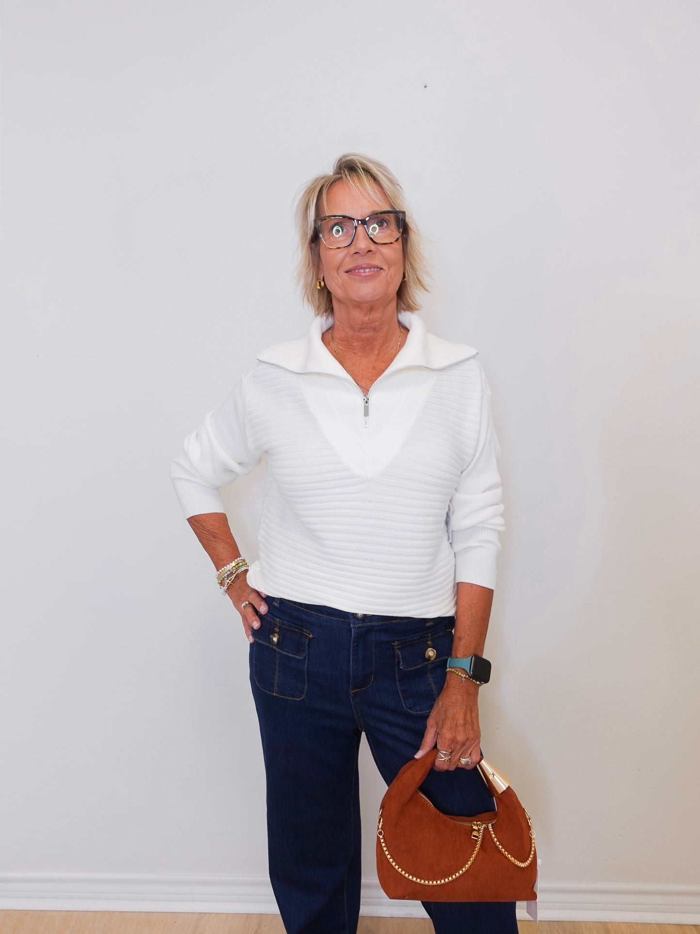 Woman wearing a white sweater and dark jeans, holding a brown handbag against a white background