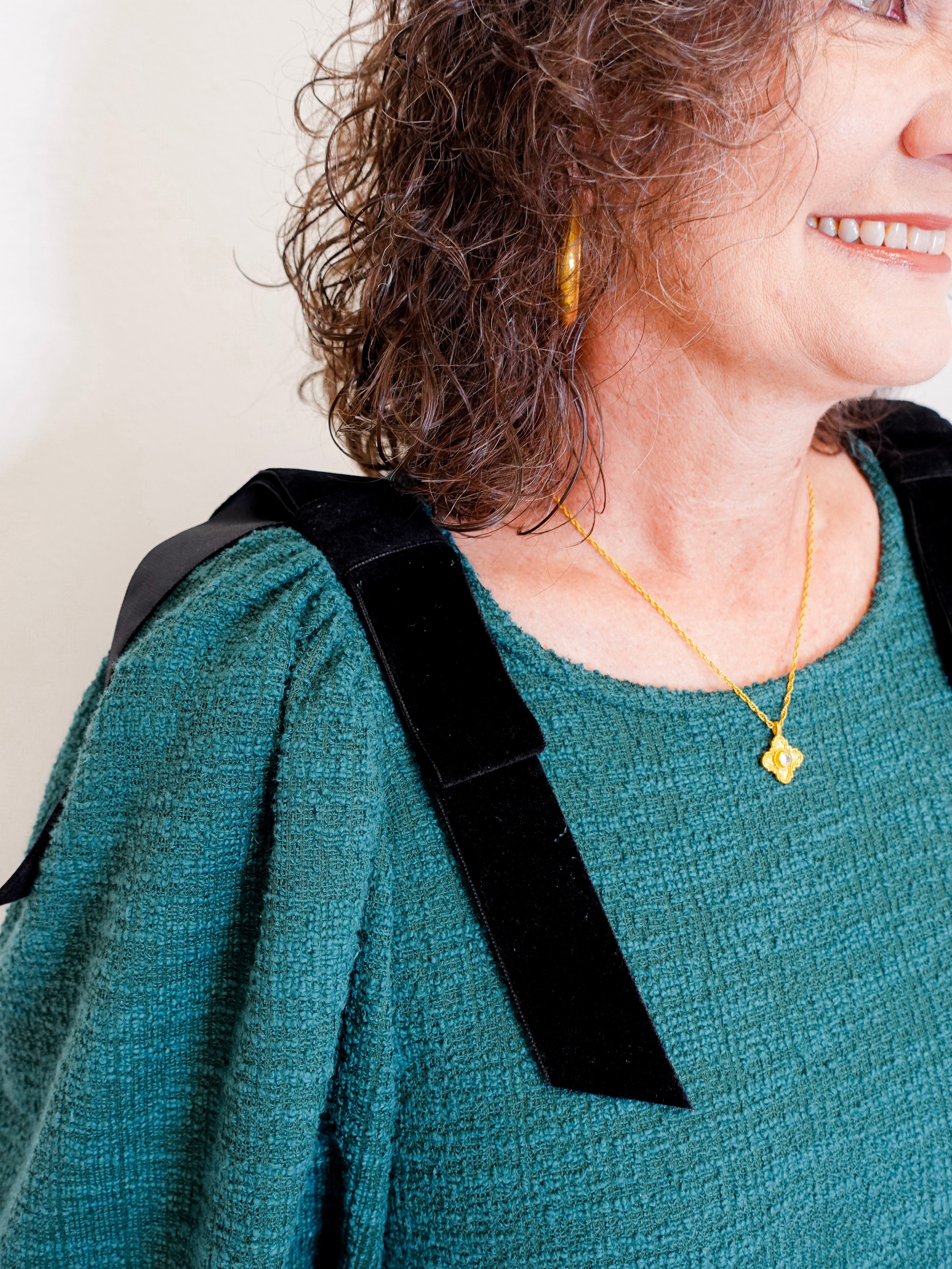Person wearing a green sweater with a black strap over one shoulder, gold necklace, and earrings.