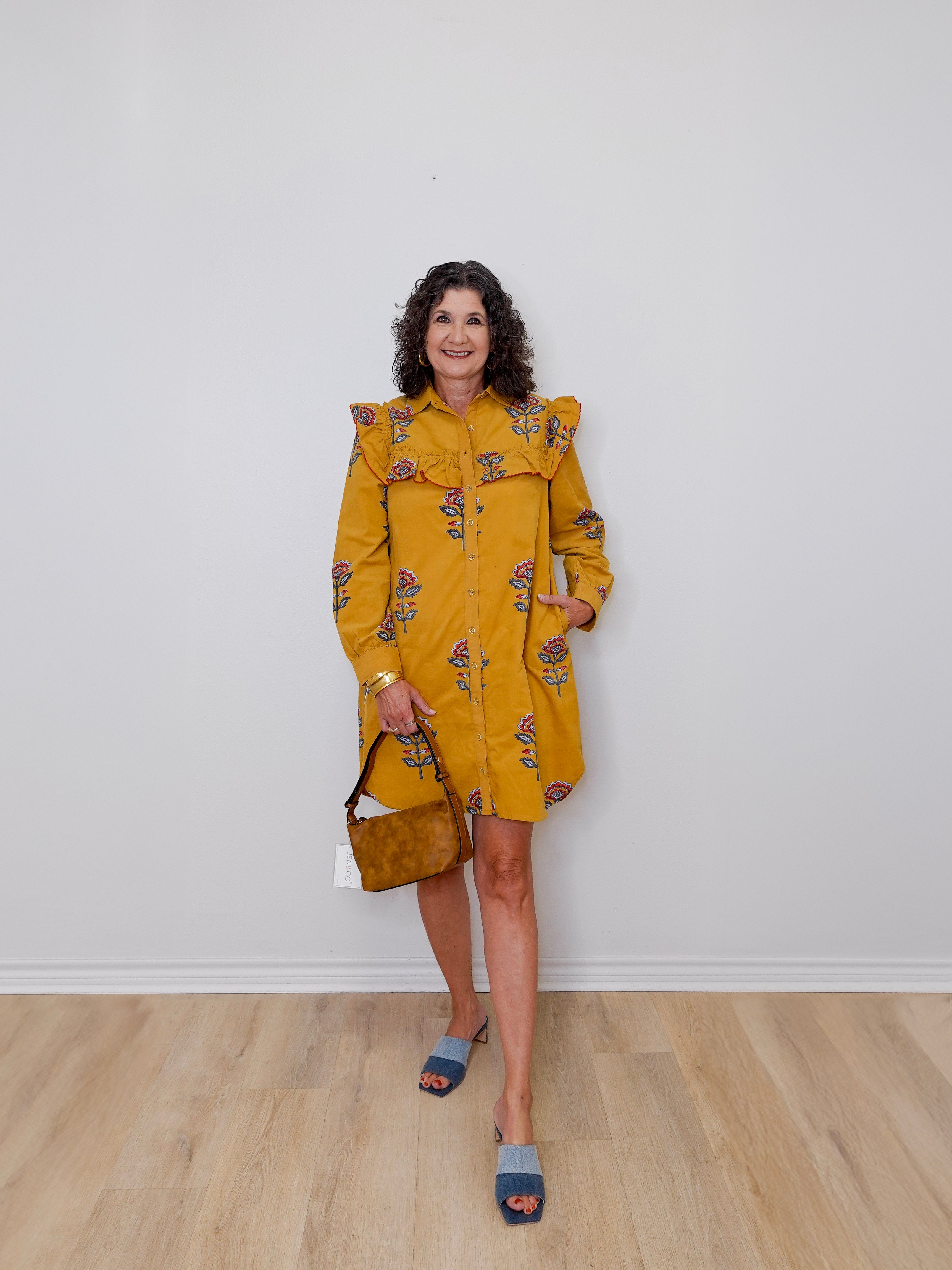 Woman wearing a mustard yellow dress with floral patterns against a white wall.