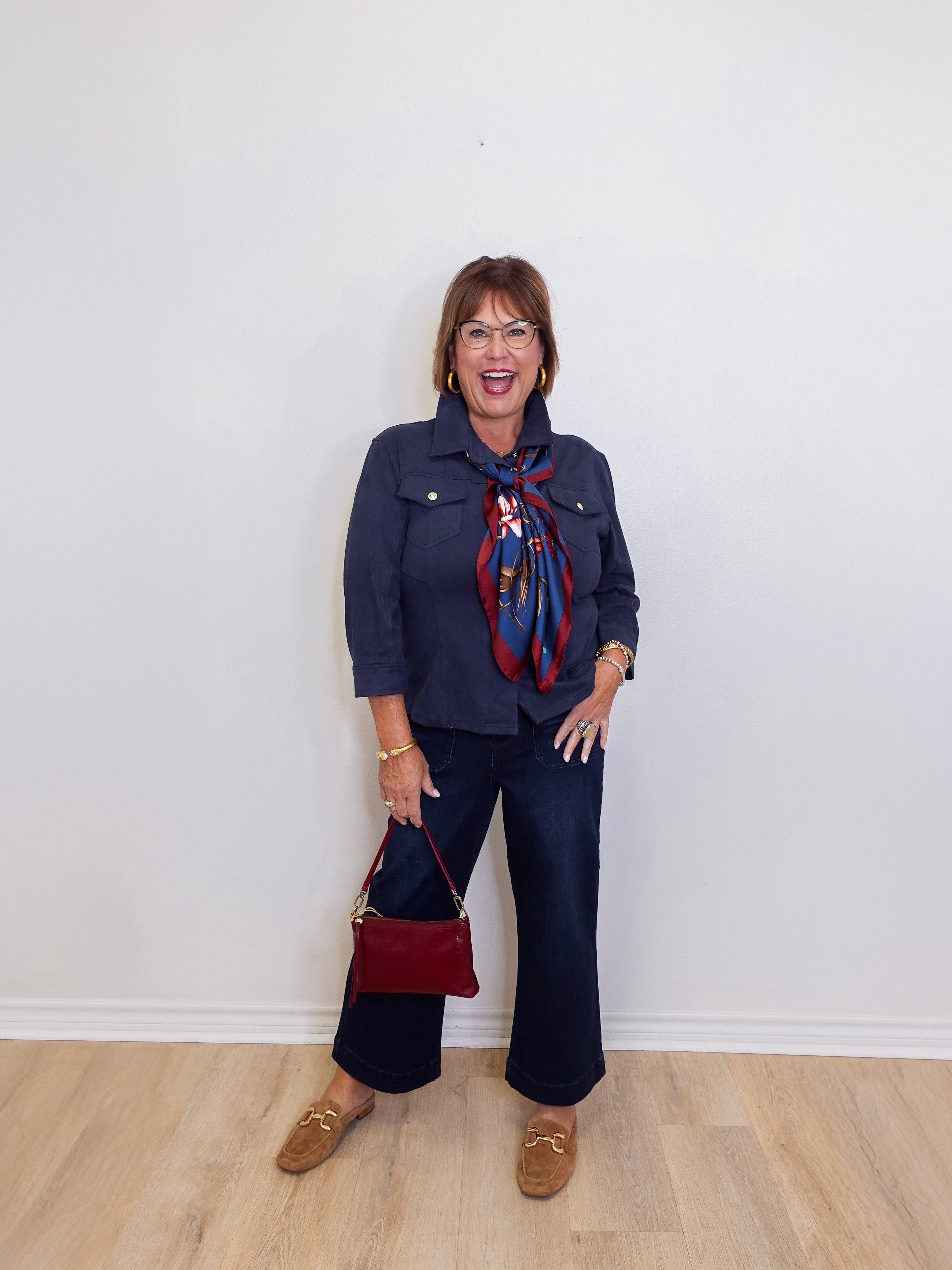 Woman wearing a navy outfit with a red bag and scarf against a white wall.