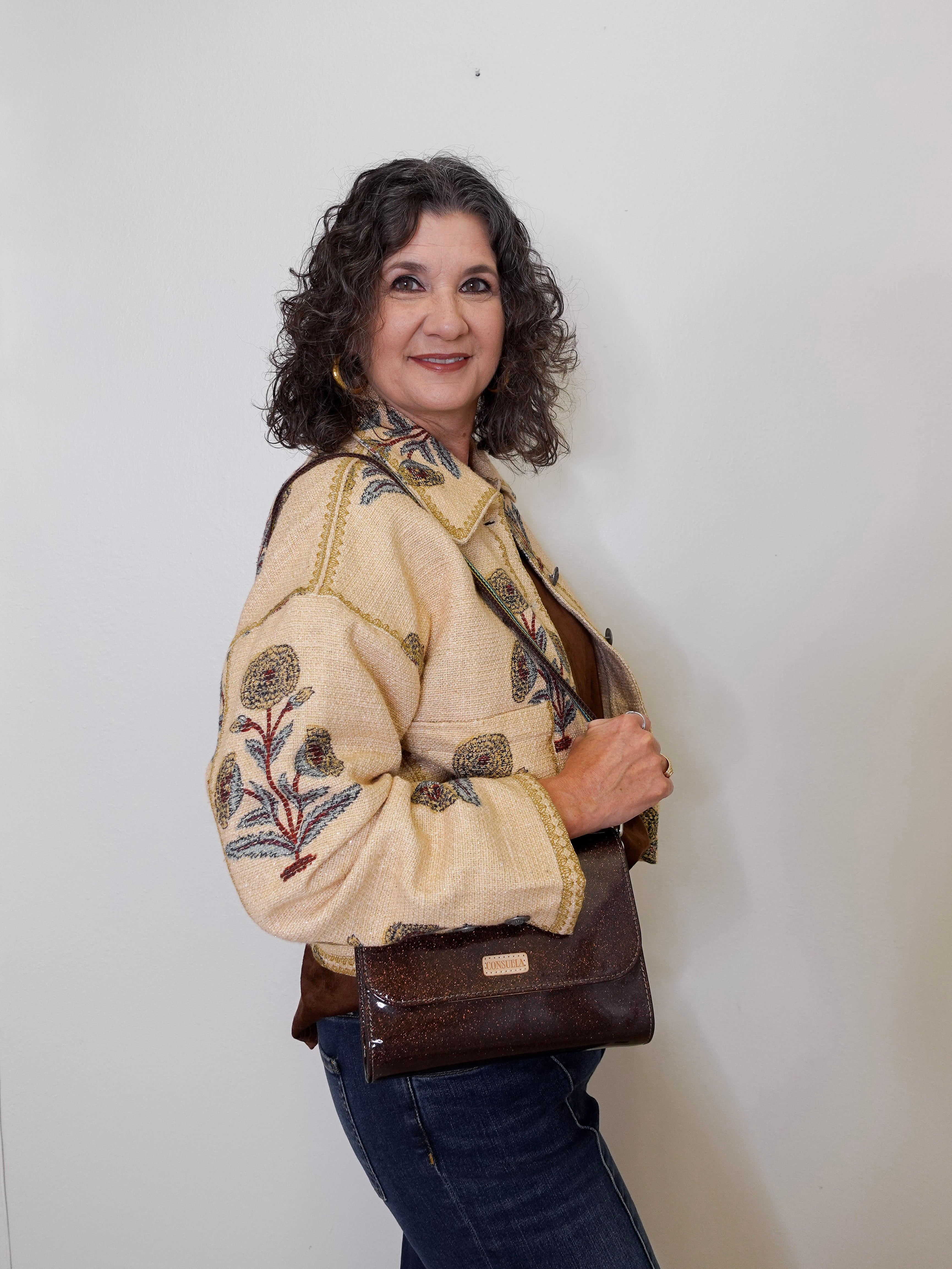 Woman wearing a gold jacket with a floral print on it, holding a brown bag against a white background.