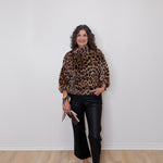 Woman wearing a leopard print top and black pants against a white wall.