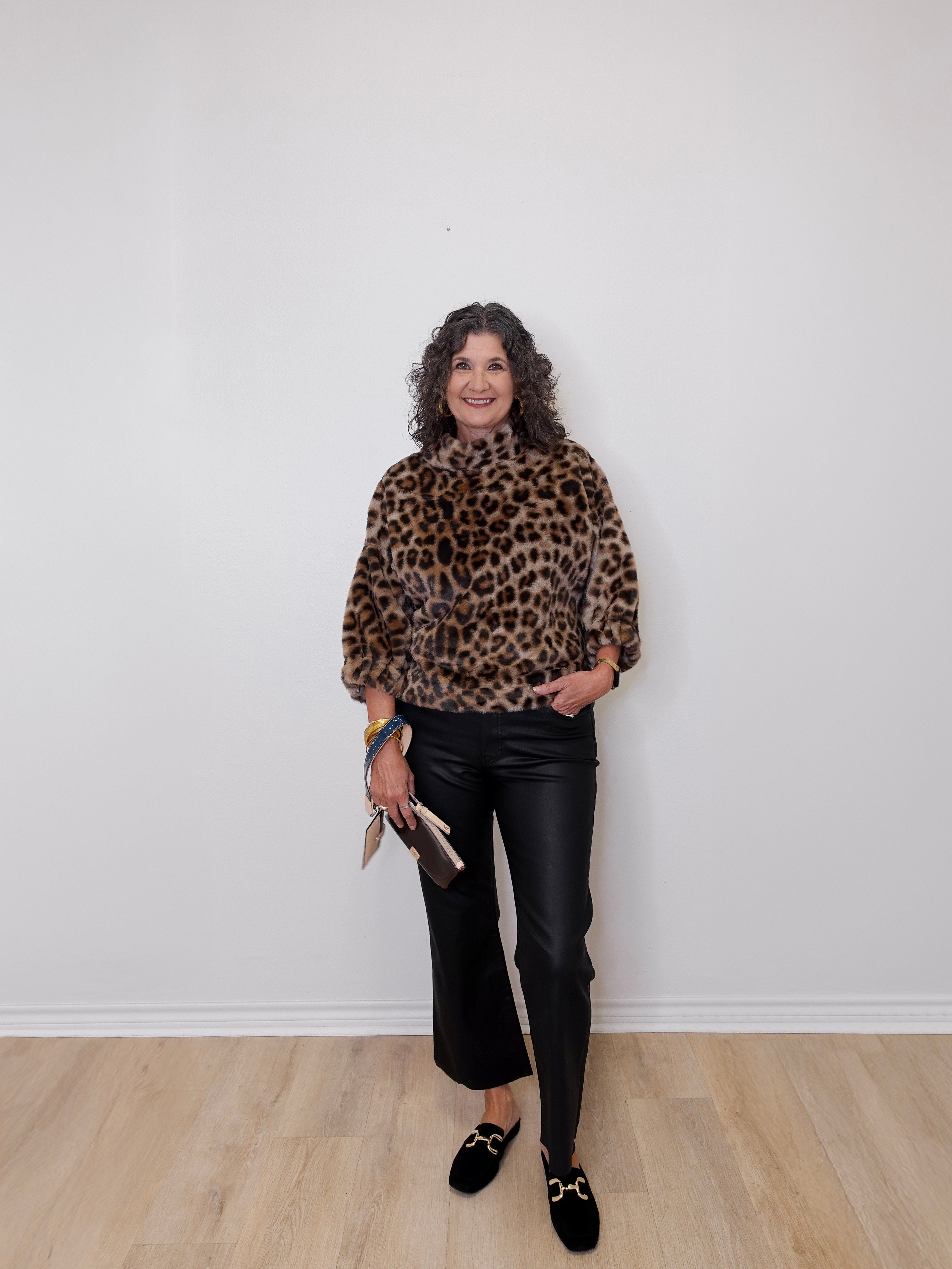 Woman wearing a leopard print top and black pants against a white wall.
