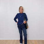 Woman wearing a blue sweater and jeans standing against a white wall.