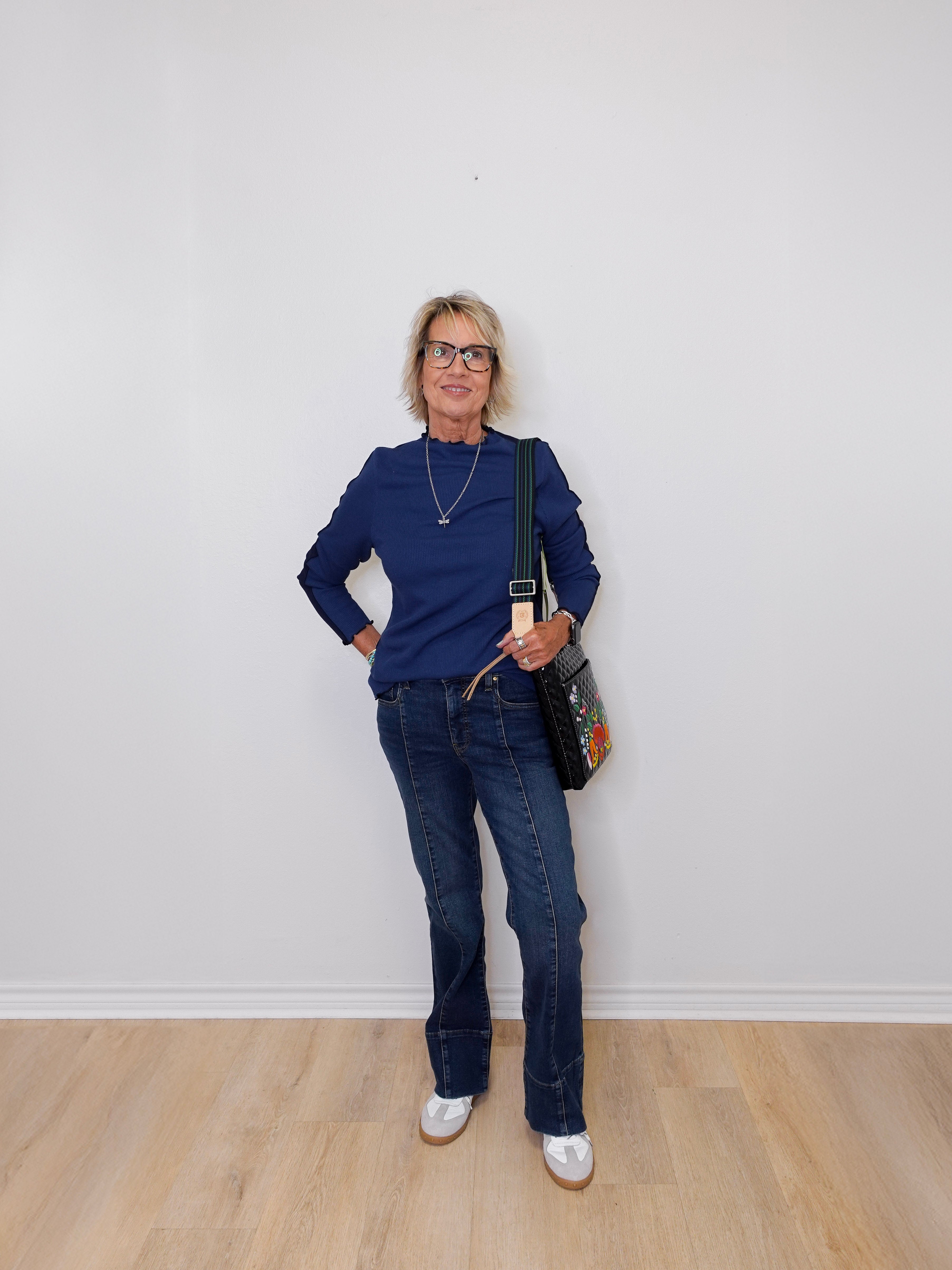 Woman wearing a blue sweater and jeans standing against a white wall.
