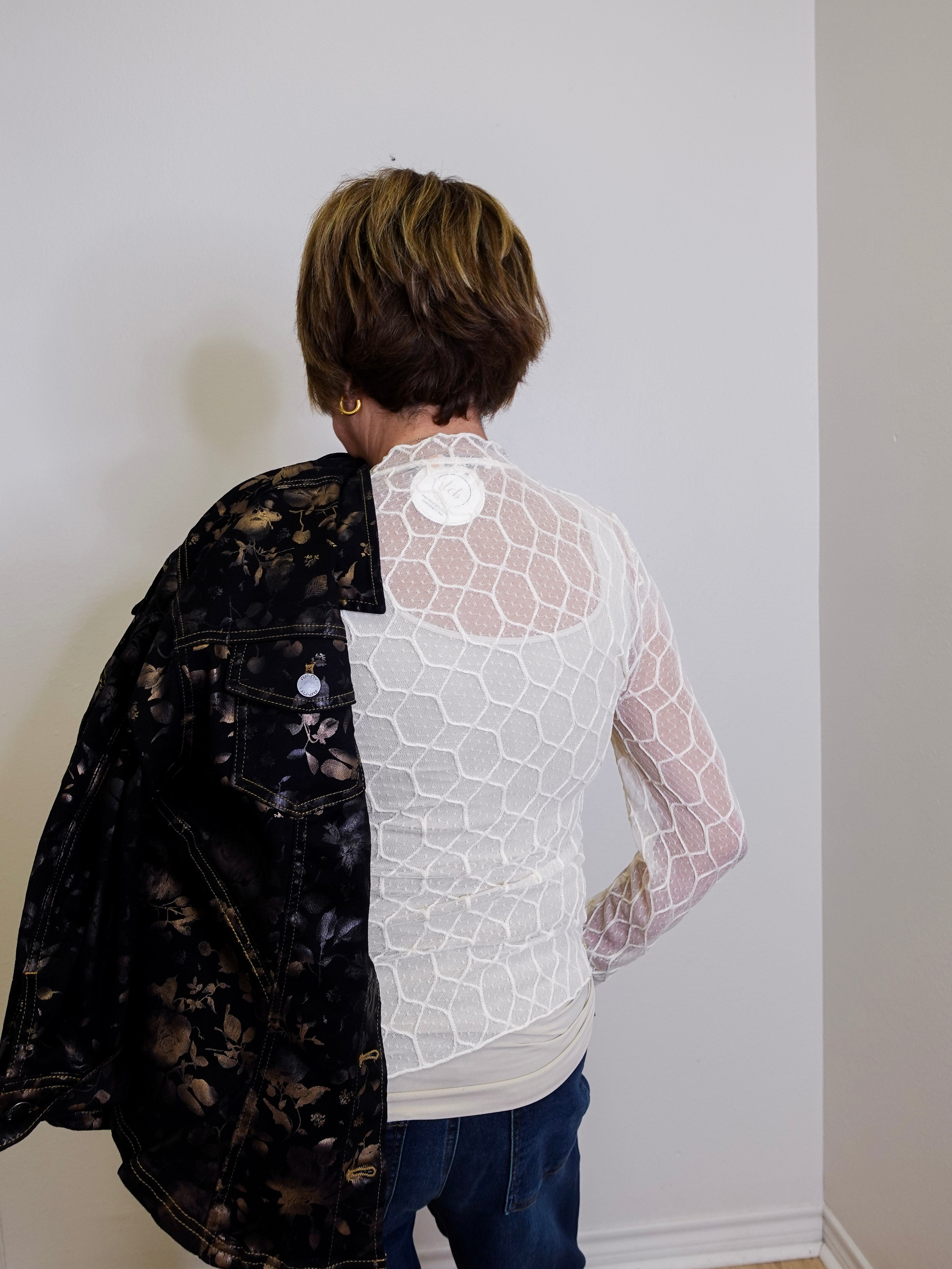 Person wearing a black floral jacket over a white lace top against a plain wall.
