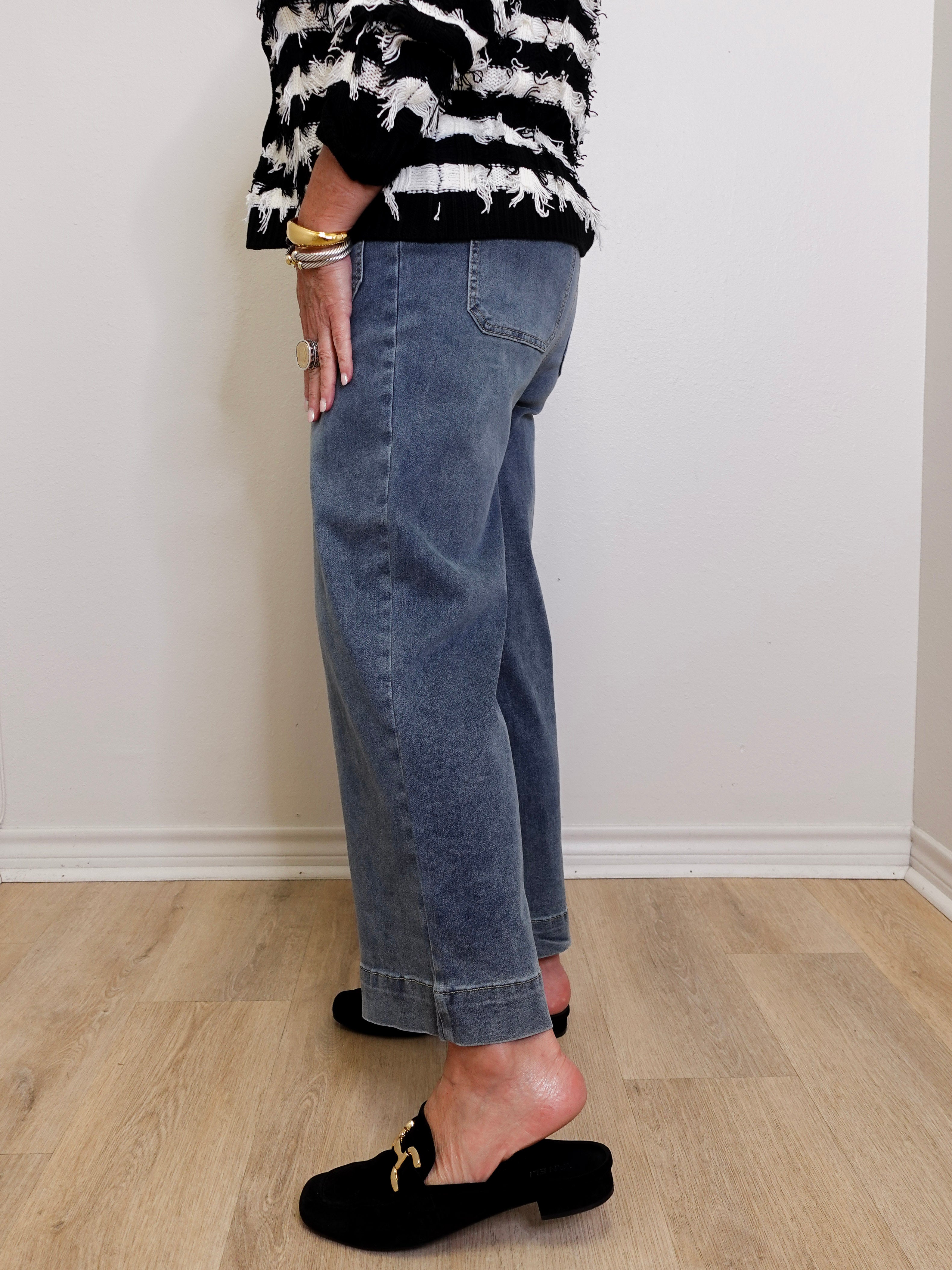 Person wearing a black and white patterned top, blue jeans, and black slippers on a light wooden floor.