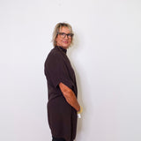 Person wearing a dark brown top and black pants against a white wall.